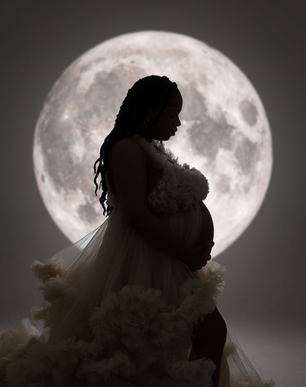 Silhouette of a pregnant woman in a ruffled white gown holding flowers, posed before a glowing full moon backdrop.