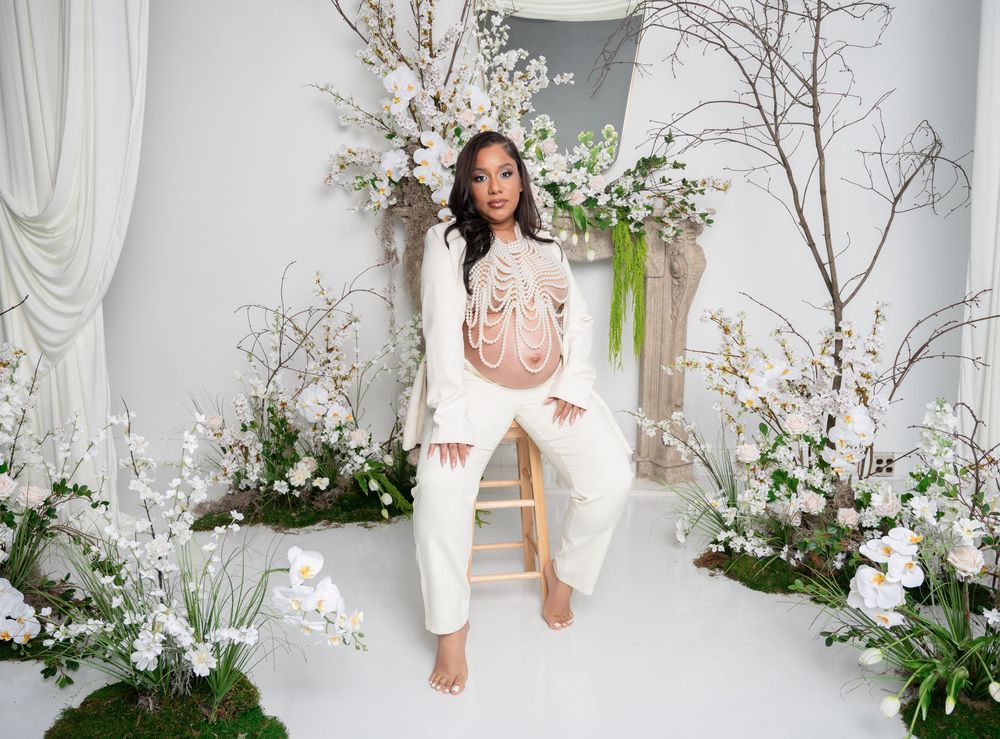 Woman in white embroidered jumpsuit seated on stool surrounded by white floral arrangements in bright studio.