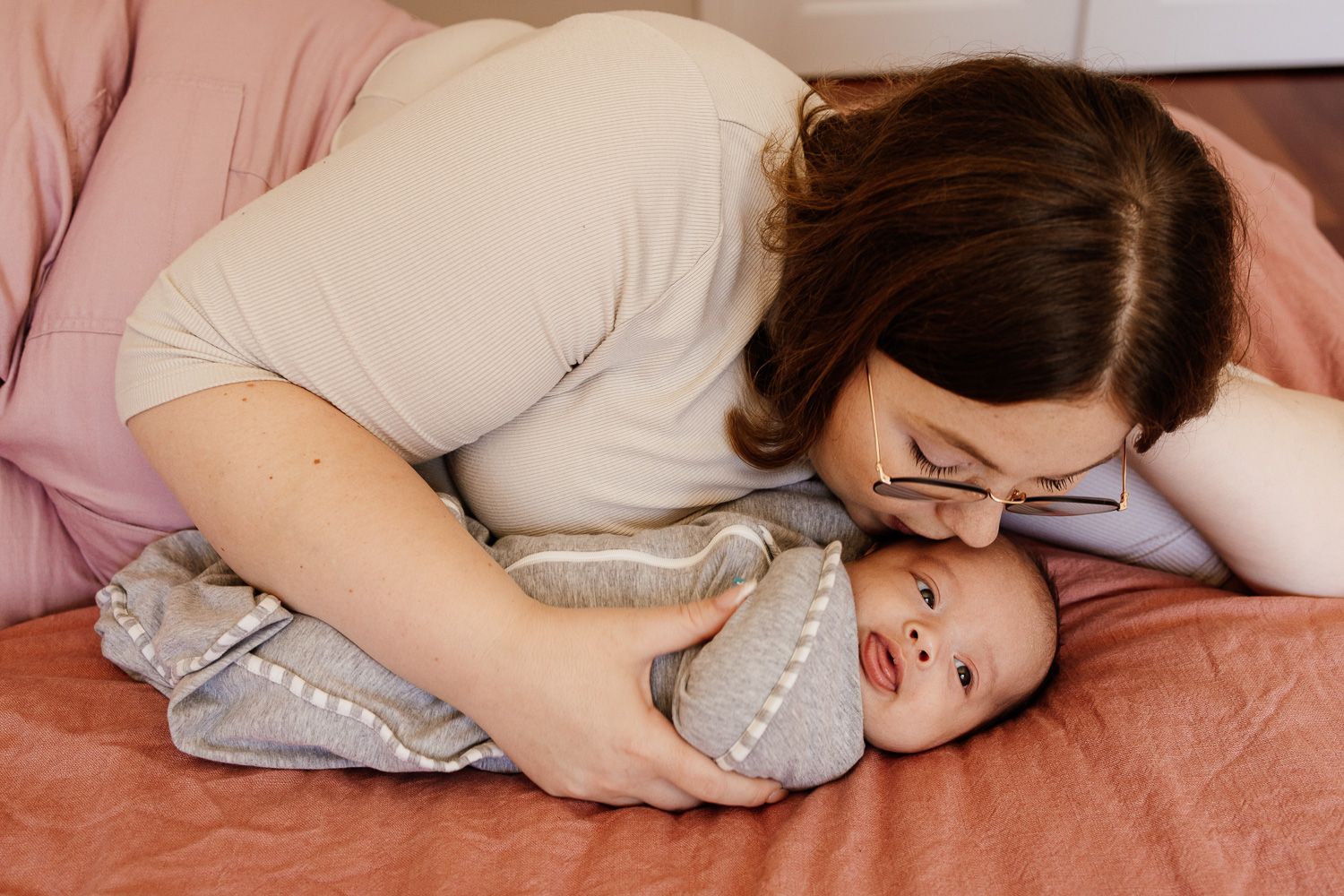 A mom in a gray sweater lying down next to her new baby, kissing him on the head