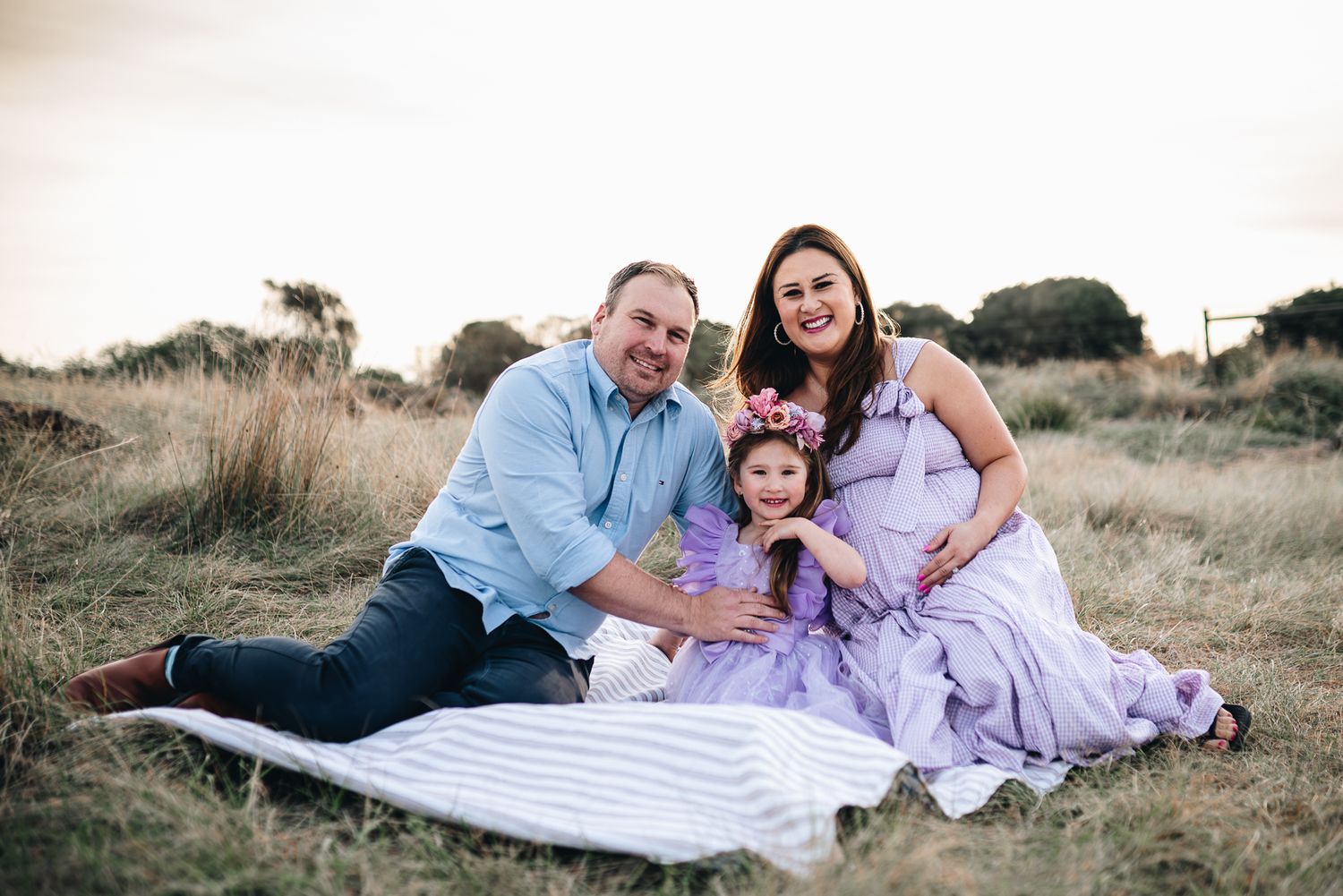 Family in coordinating purple and blue outfits poses together on grassy hilltop during golden hour taken by Melbourne maternity photographer
