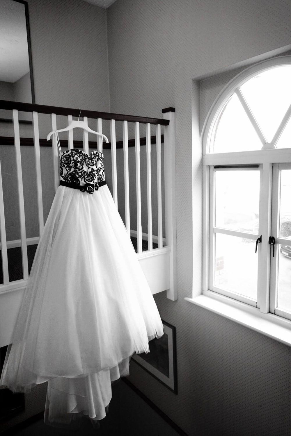 A white wedding dress hangs elegantly near windows and white railings in a bright interior space.