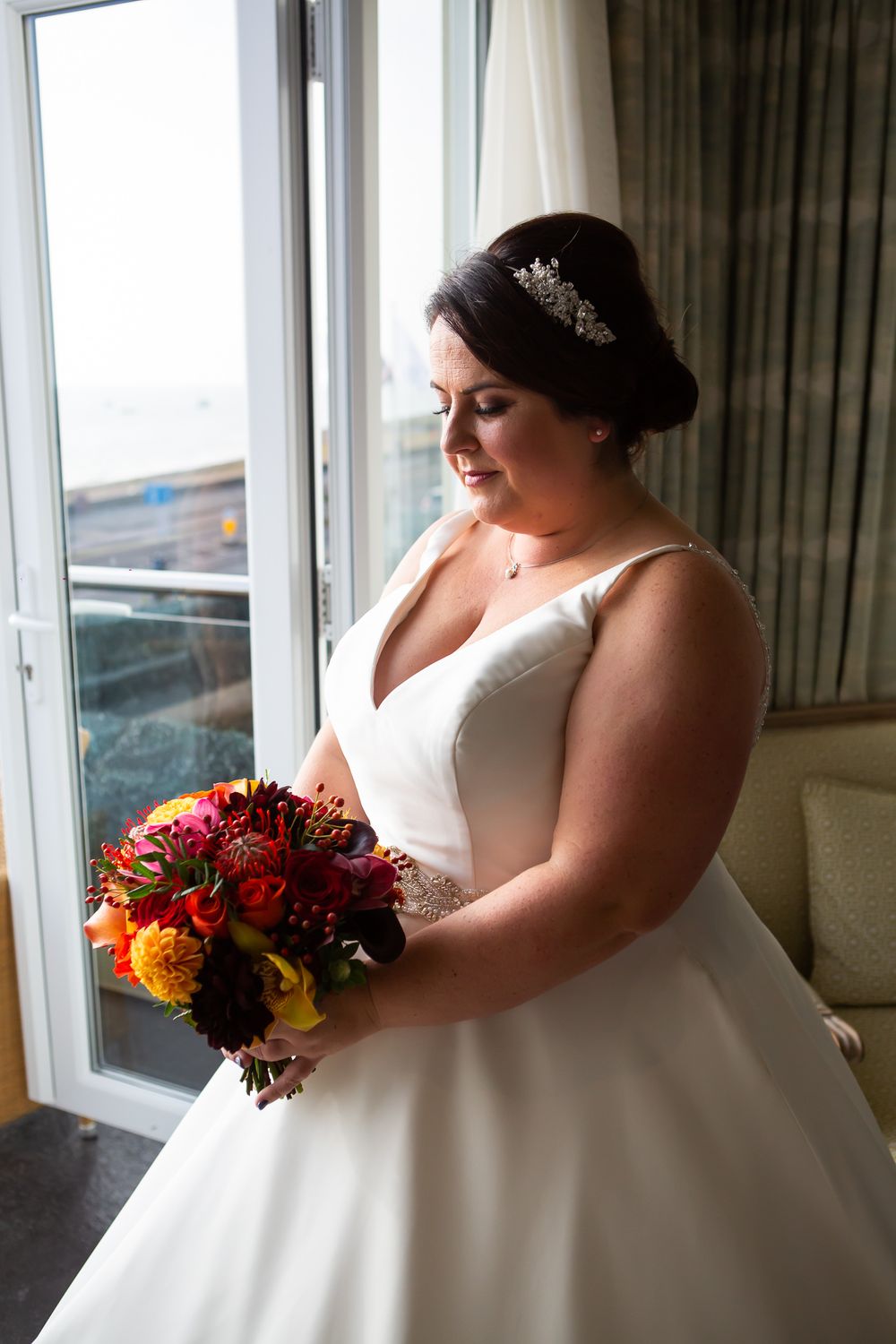 A bride holding a vibrant autumn bouquet with orange and red flowers stands by a window.