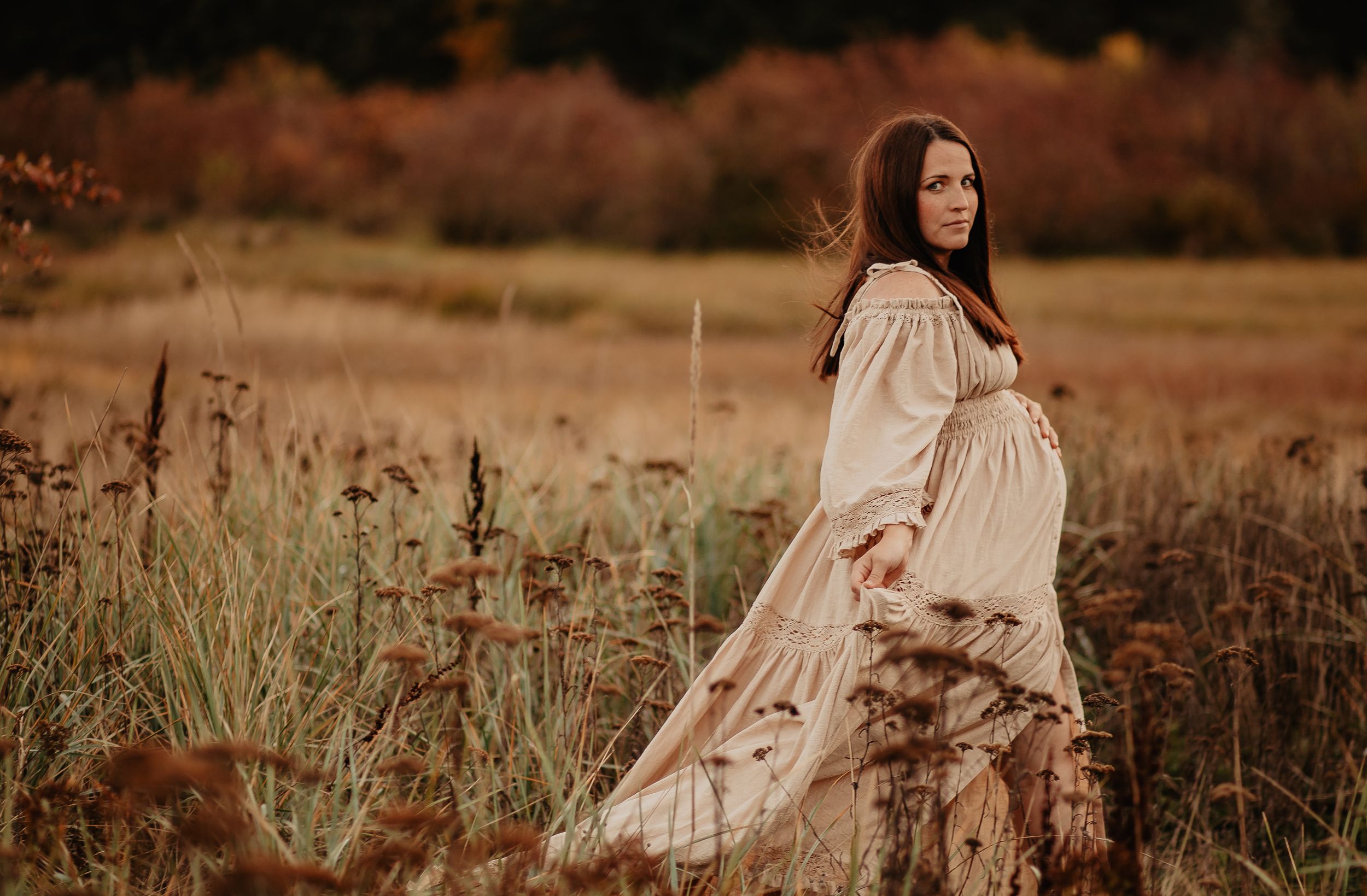 A woman in a flowing white dress stands gracefully in a golden autumn field during sunset.