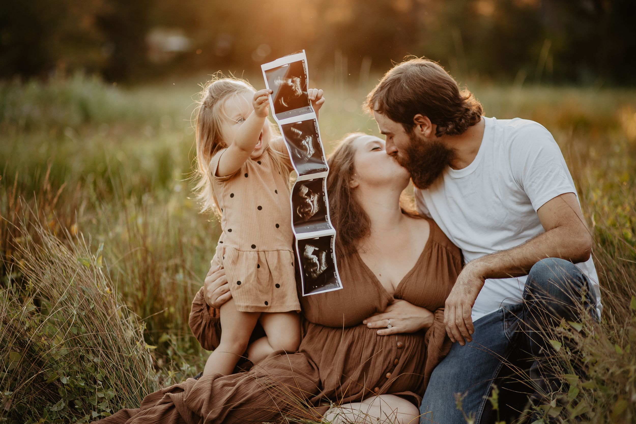 A couple sits together in a sunlit meadow holding ultrasound photos while sharing an intimate moment at sunset.