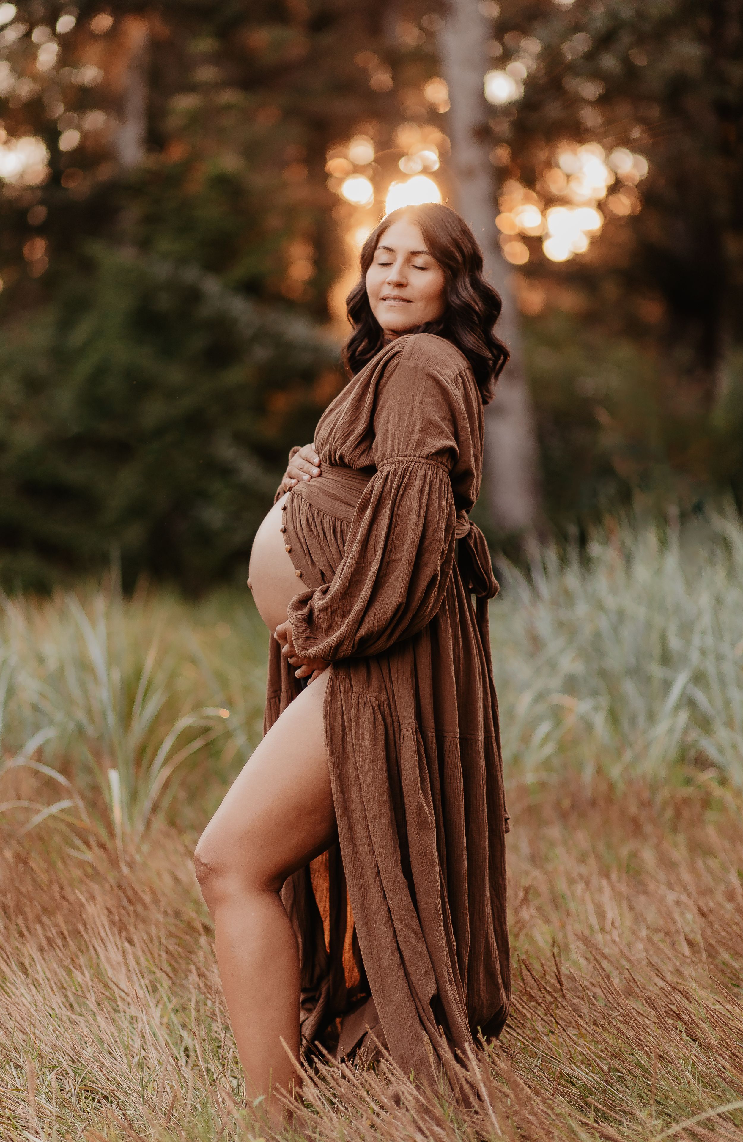 Pregnant woman in a brown robe in a field at golden hour embracing her unborn child
