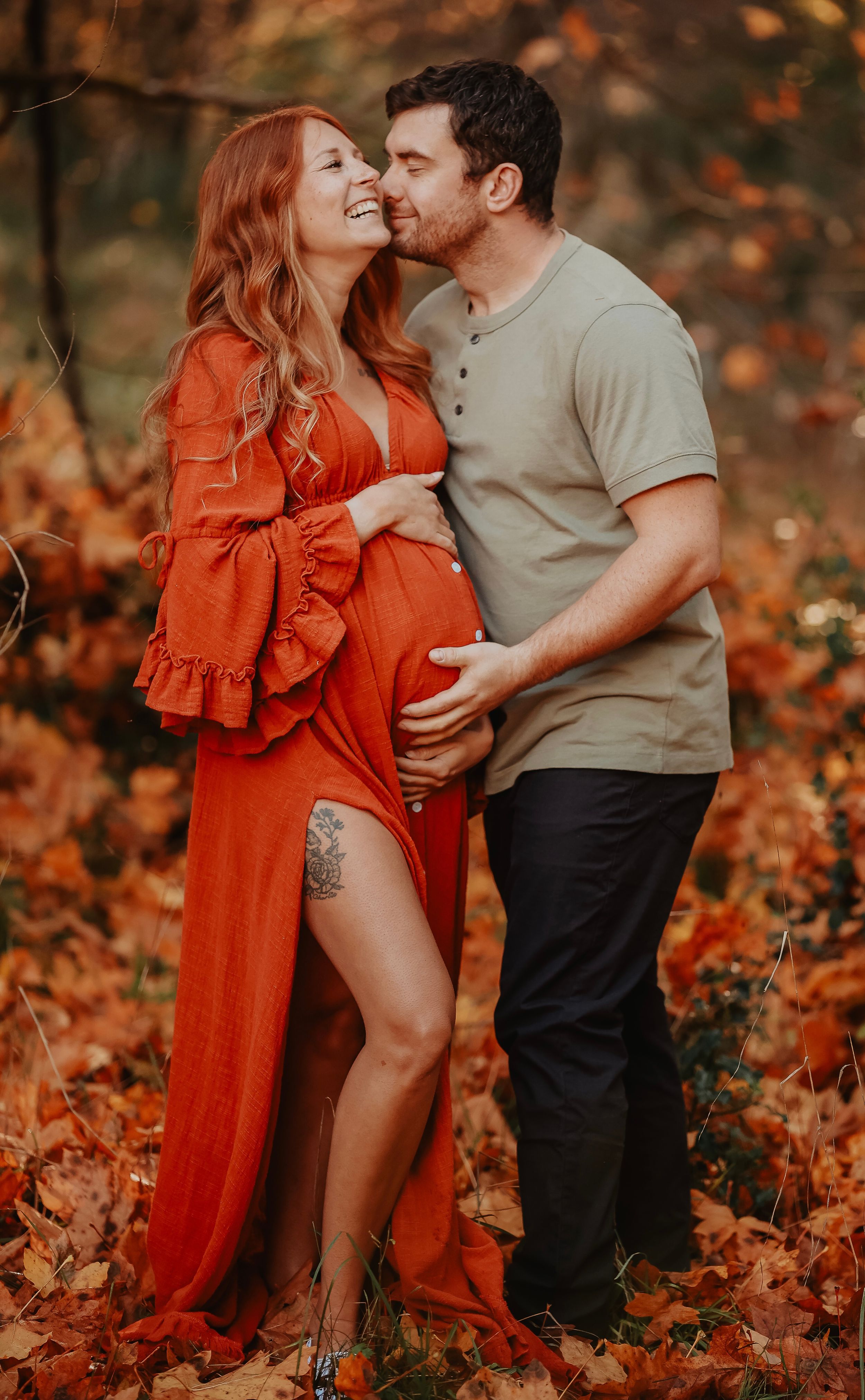 A couple embraces intimately among fallen autumn leaves as the woman wears a flowing red dress in a romantic outdoor setting.