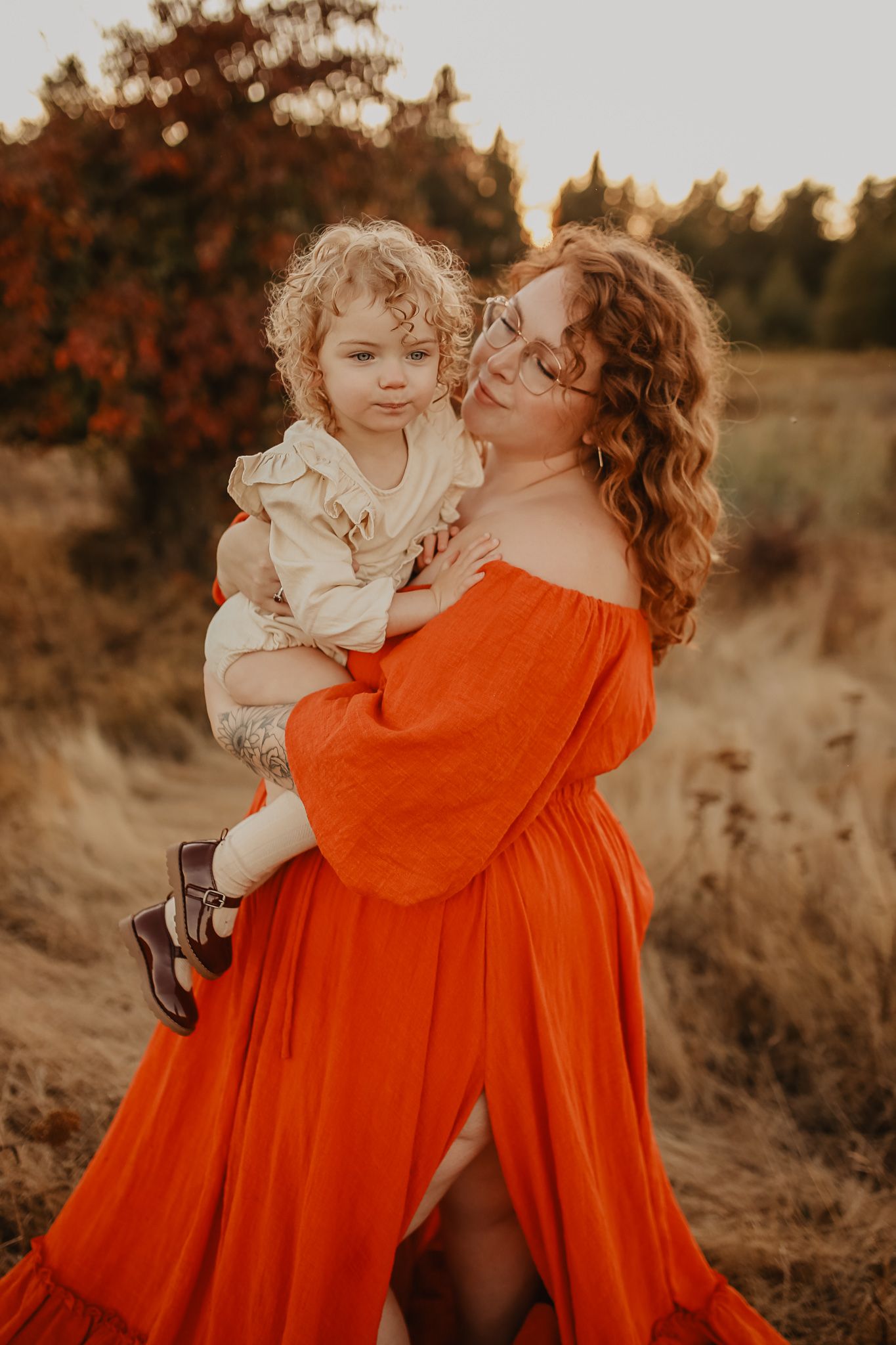 A parent in a flowing orange dress holds and embraces a small child in a white dress during a golden hour photoshoot.