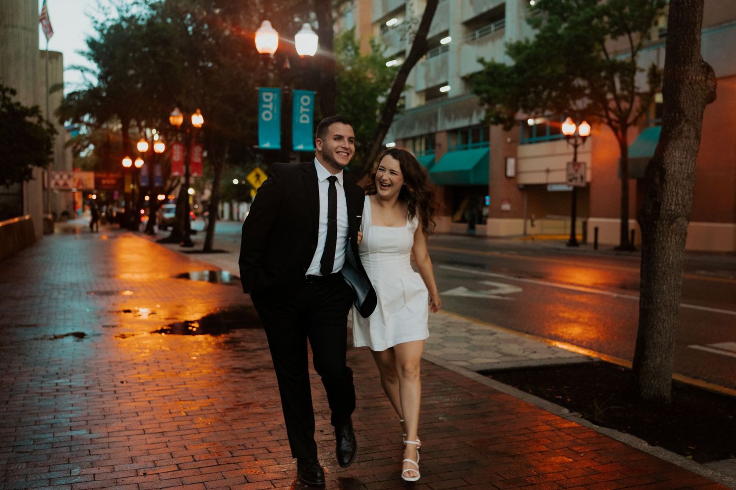 A couple walks down a city street at dusk with glowing street lamps creating a romantic urban atmosphere.