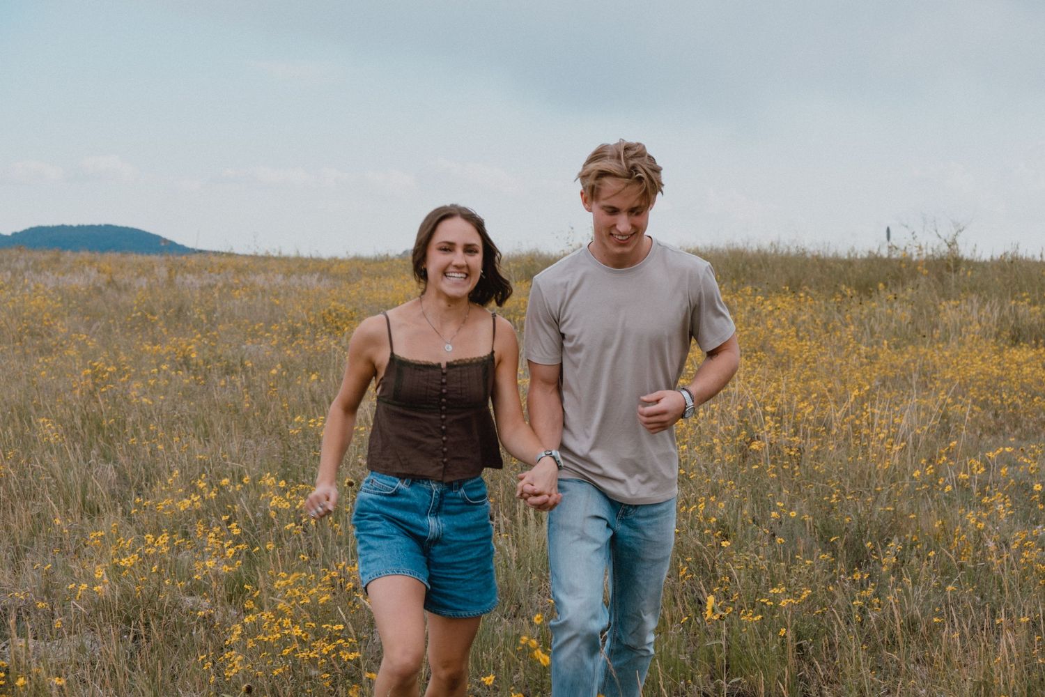 Two people running and laughing together through a scenic mountain meadow filled with wildflowers.