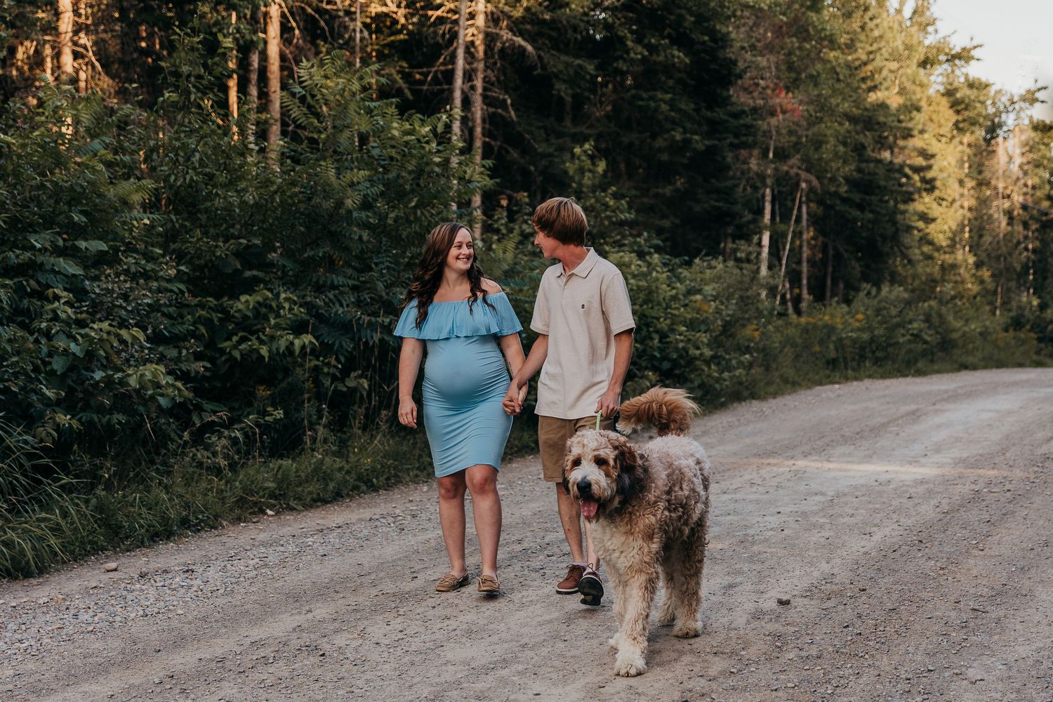 Two people walking with a large dog on a dirt road surrounded by green forest at sunset celebrating their new chapter of pregnancy.