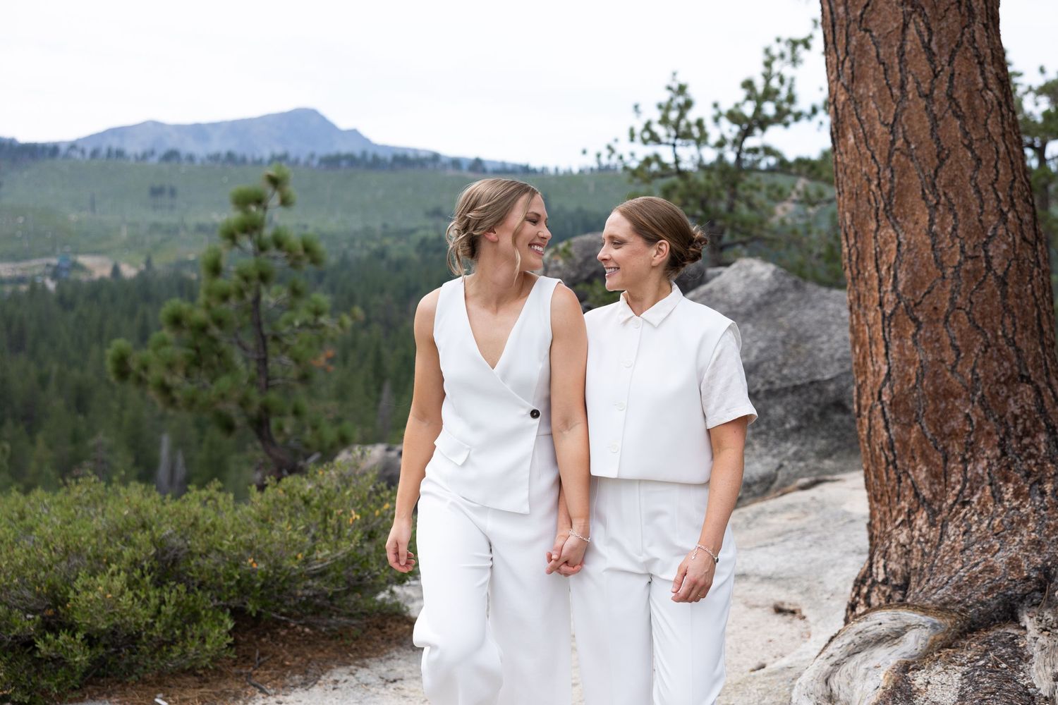 Two women in white outfits hold hands and smile while walking along a scenic mountain trail.