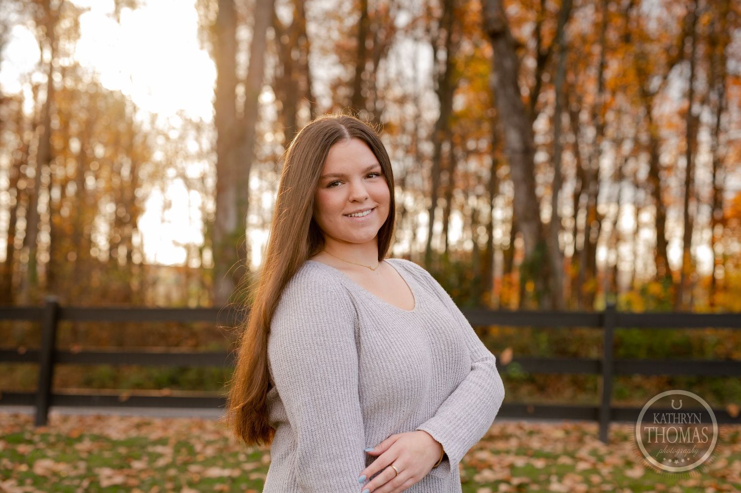 Portrait of someone in a cozy sweater against a backdrop of autumn trees and wooden fencing.