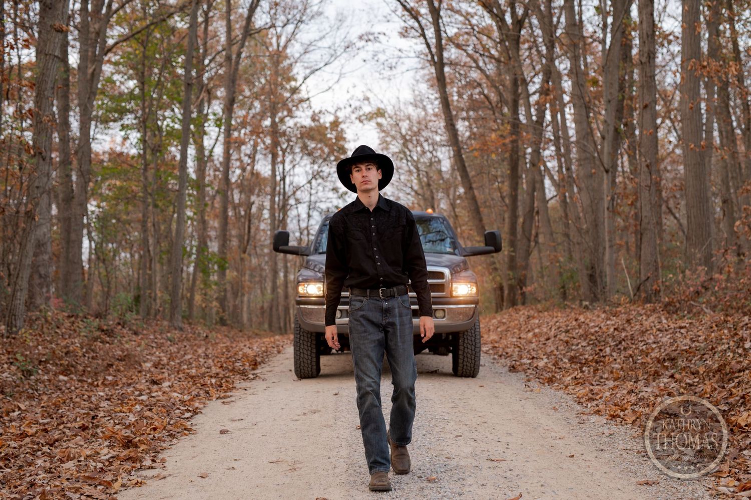A figure wearing a black hat walks on a leaf-covered path with a truck's headlights visible behind.