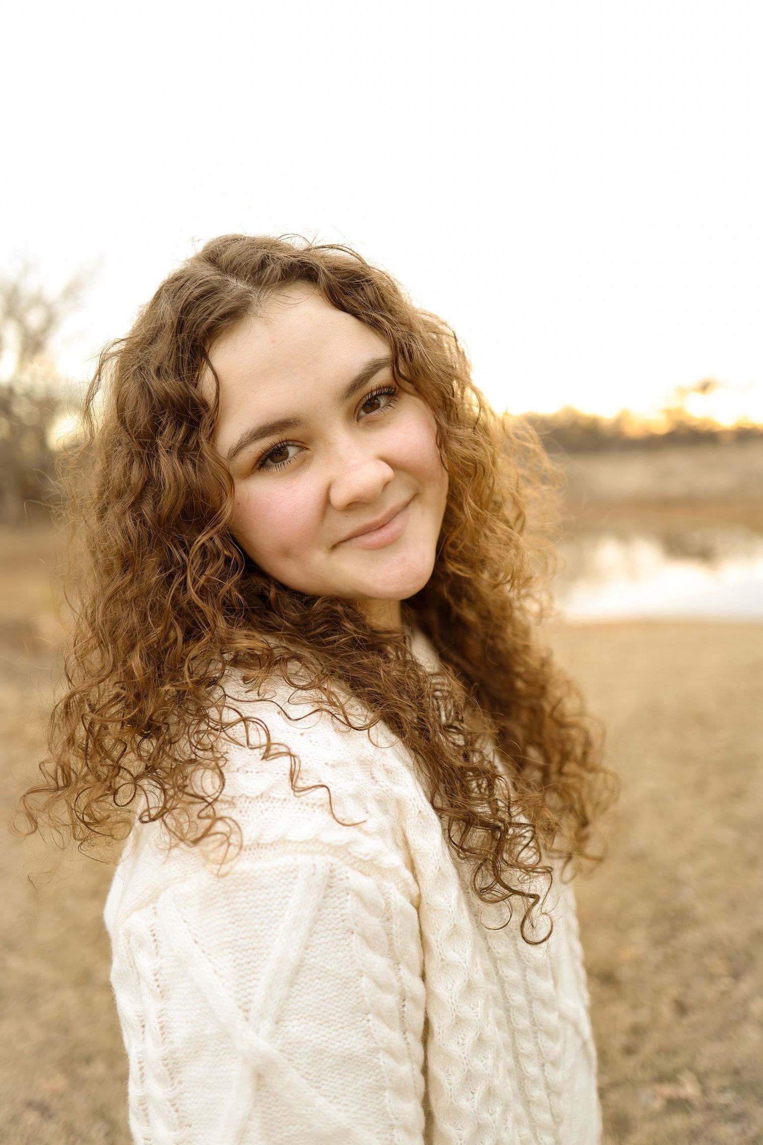 Portrait of someone with long curly hair wearing a white top in warm sunset lighting outdoors.