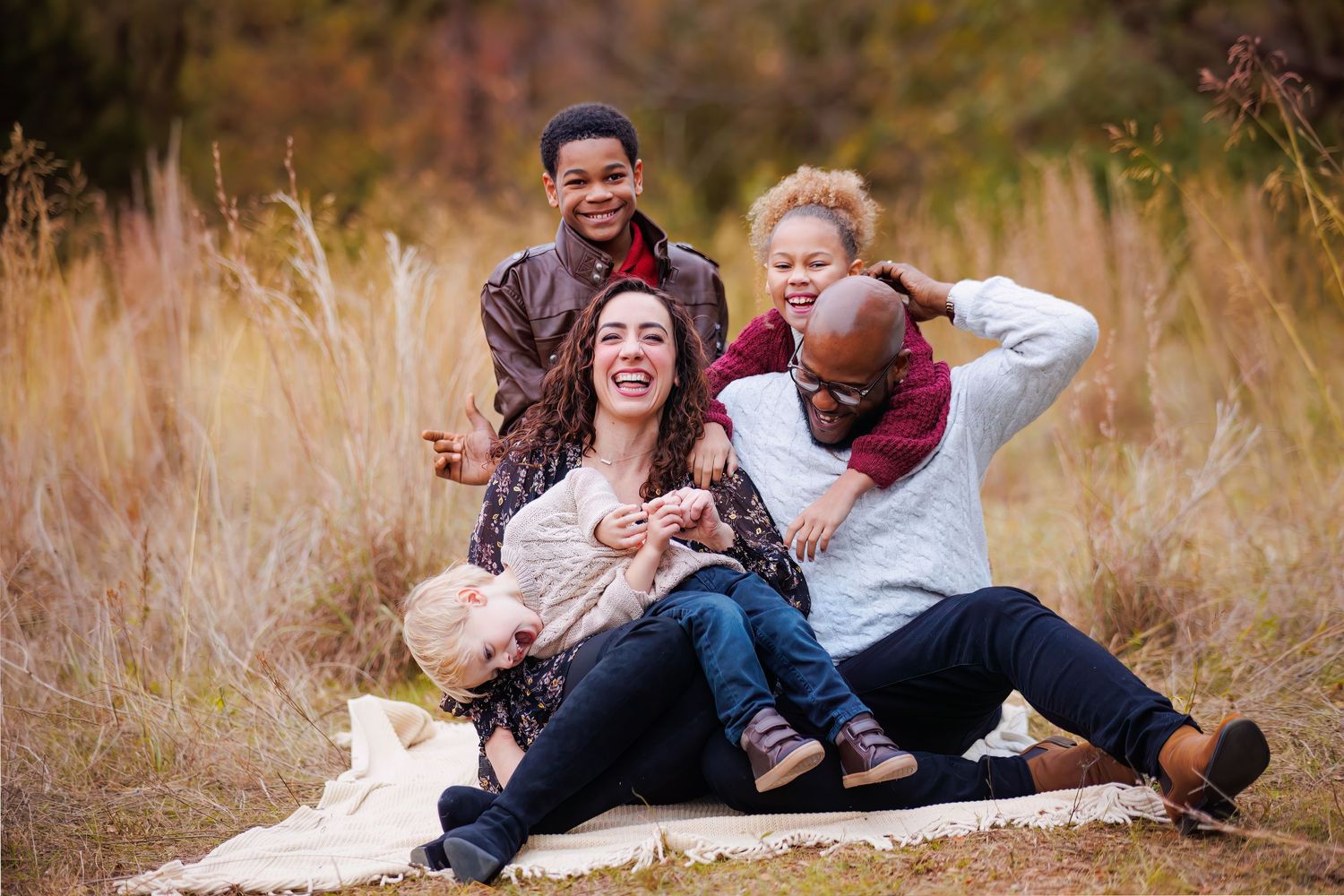 Family laughing together while sitting on blanket in autumn field with tall grass.