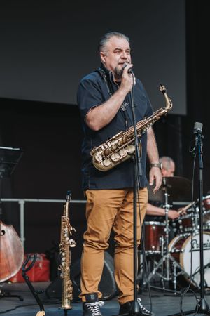 Saxophone player performs on stage with drum kit visible in background.