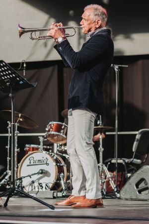 Trumpet player in navy blazer and light pants performs on stage with a drum kit visible behind.