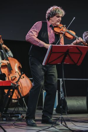 Violinist in striped vest performs on stage with a music stand and cello visible in background.