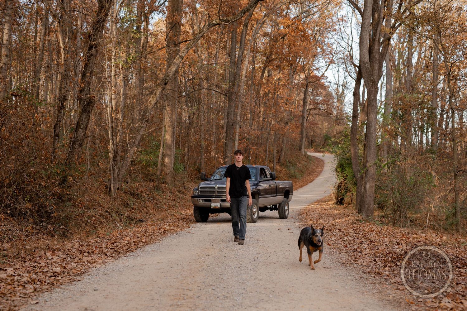 Person walking with dog on dirt path through fall forest with truck in background.