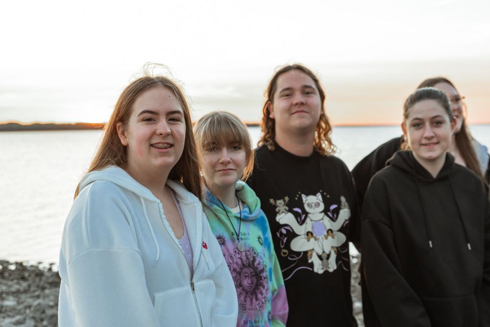 A group of four friends in casual attire standing together by the water at sunset.