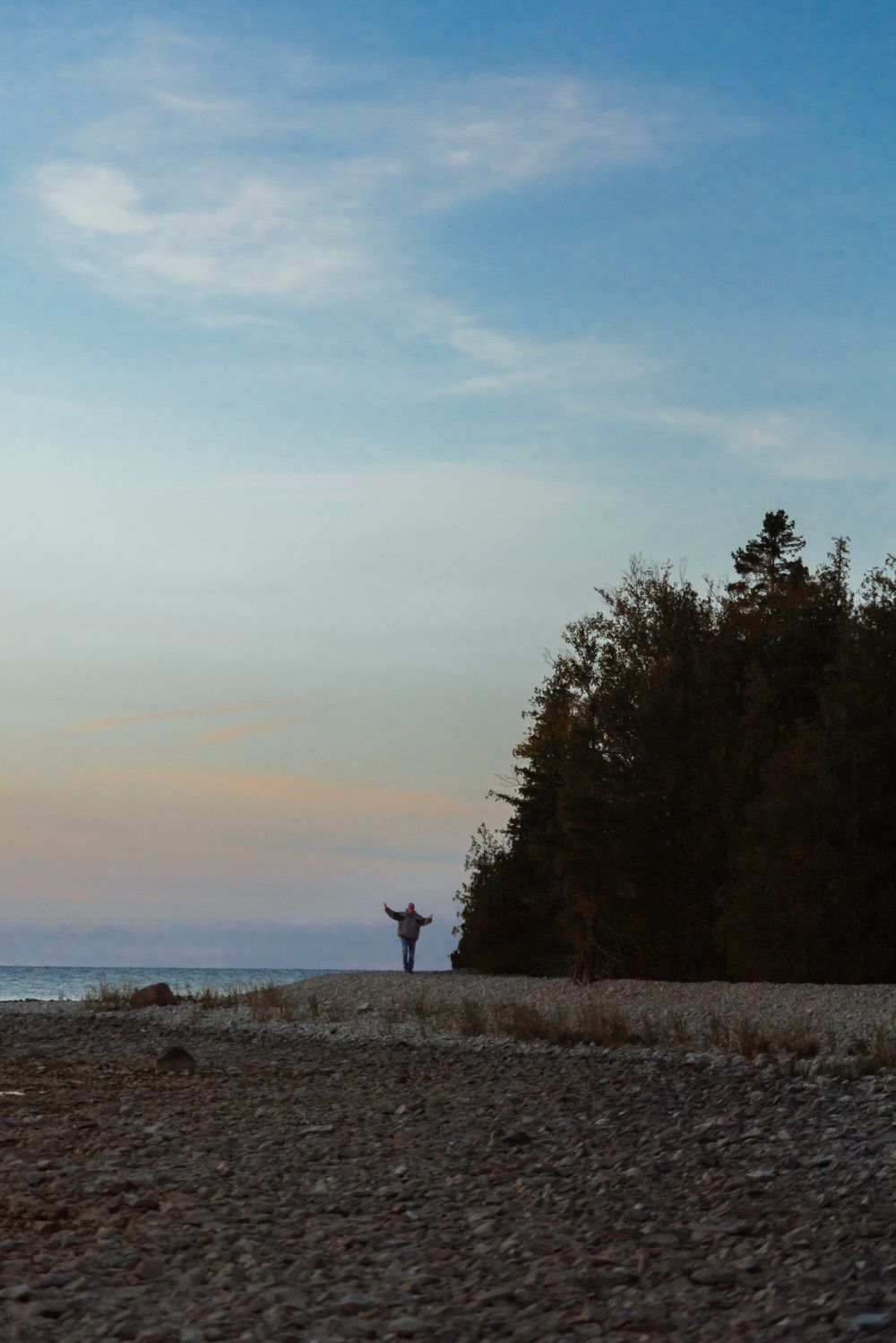Silhouette of a person with raised arms stands on a rocky beach at sunset near a forested coastline.