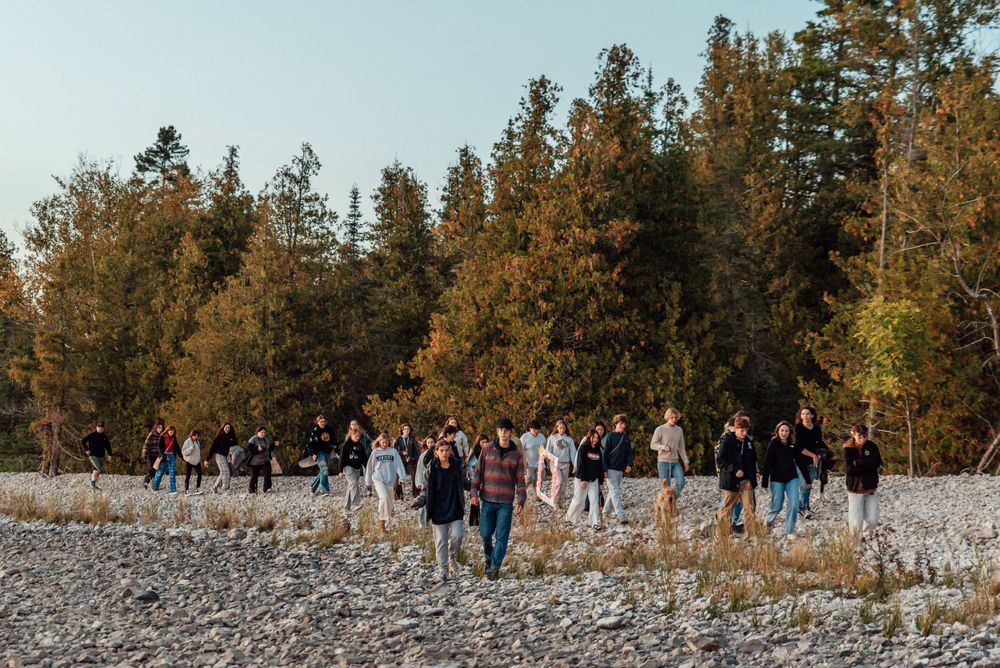 Group of people walking along a rocky beach path bordered by autumn trees.