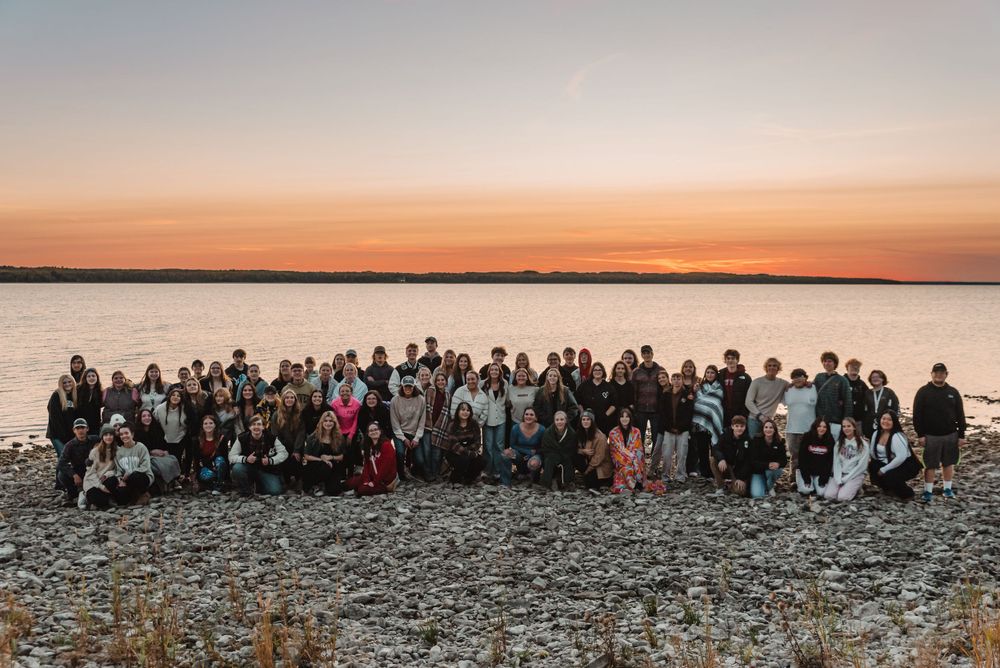 Large group gathered on a rocky shoreline during golden hour sunset.