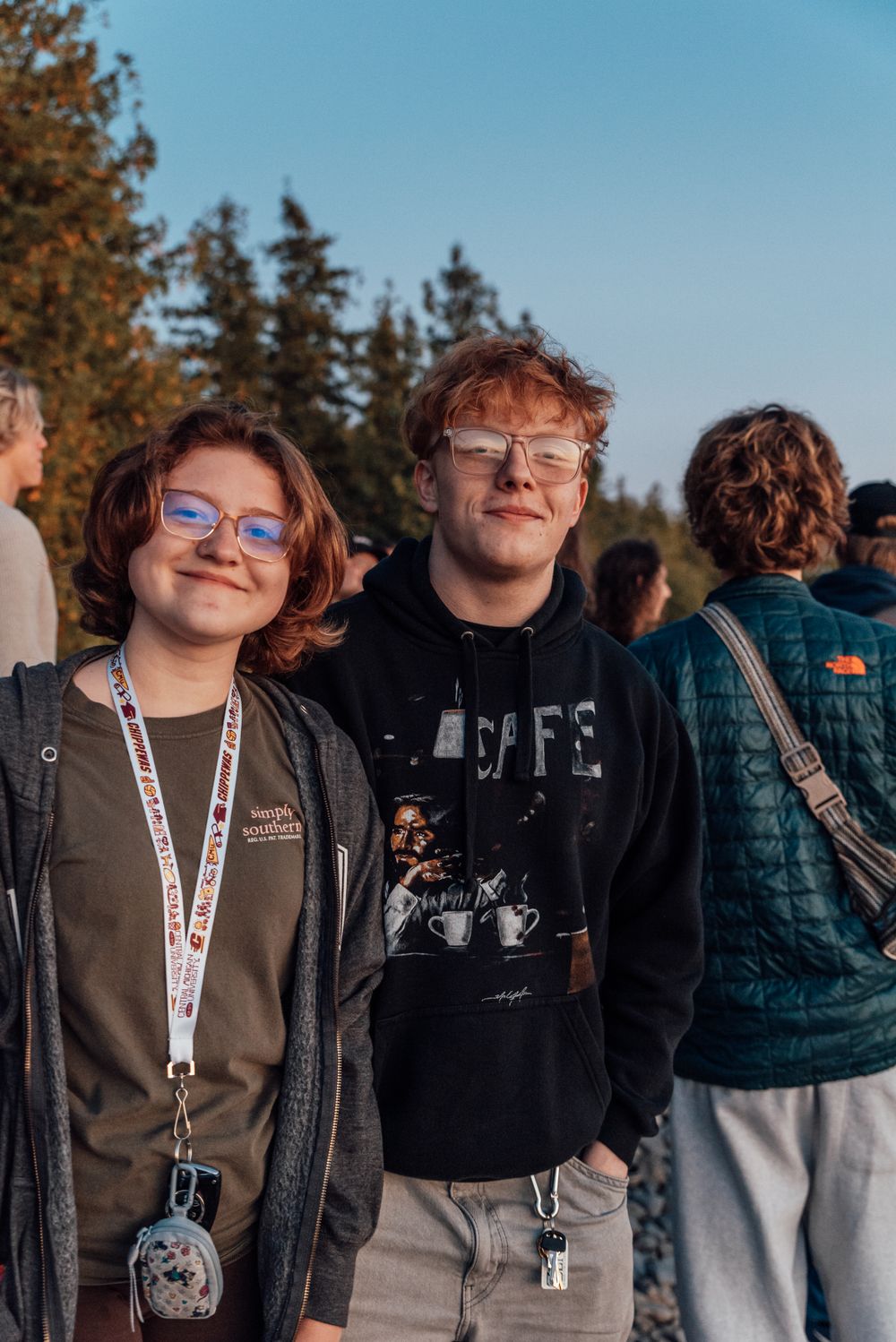 Two students wearing casual attire and lanyards smile together outdoors with trees in the background.