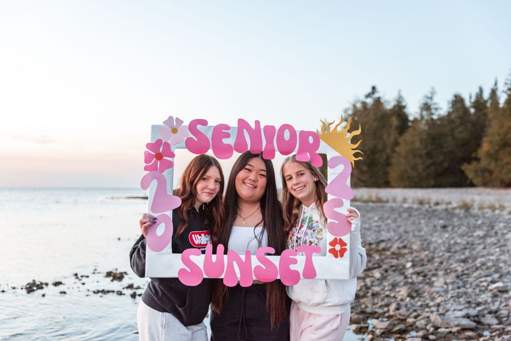 Three students pose with a pink senior photo frame prop on a rocky beach at sunset.
