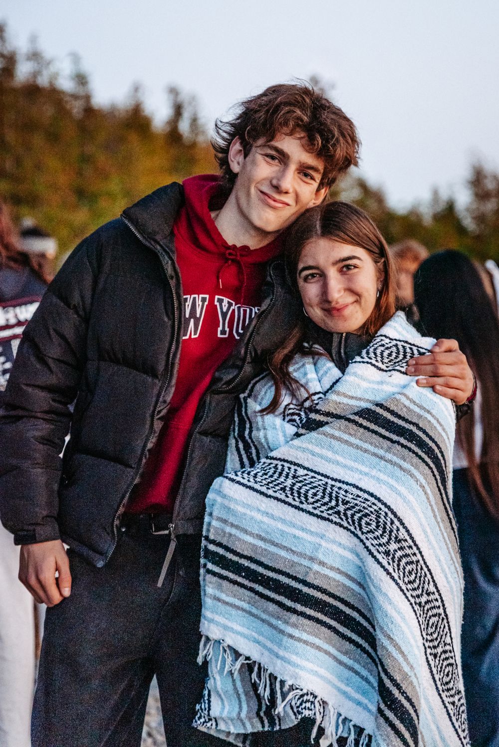 A couple stands together outdoors, with one wearing a puffer jacket and the other wrapped in a patterned blanket.