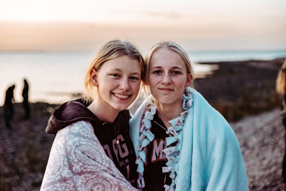 Two friends share a moment together on a rocky beach during sunset, wrapped in cozy blankets.