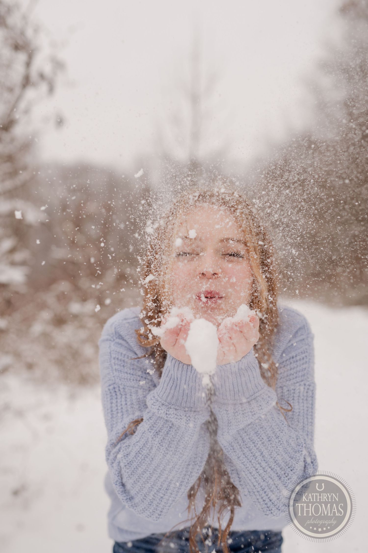 A person in a blue sweater blows snow into the air during a winter photoshoot in a snowy forest.