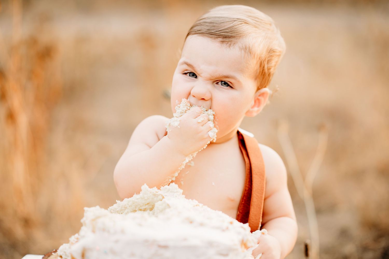A baby in brown suspenders sits in a field eating cake with frosting smeared on their face during a golden hour photoshoot in Puyallup.