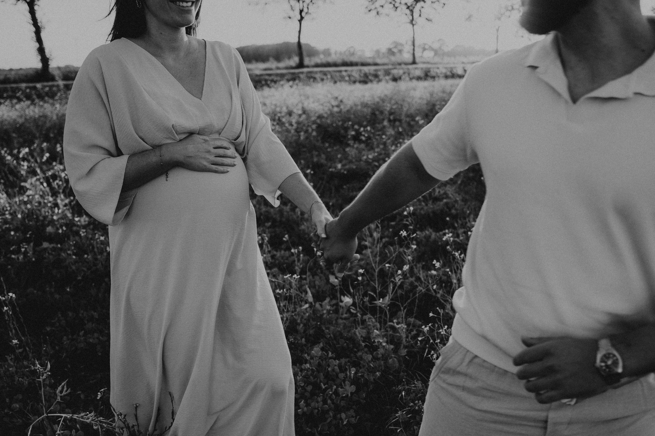 Man en vrouw in lichte kleding. Vrouw is zwanger. Lopen hand in hand door een veld met gras en bloemen. Zwart wit foto.