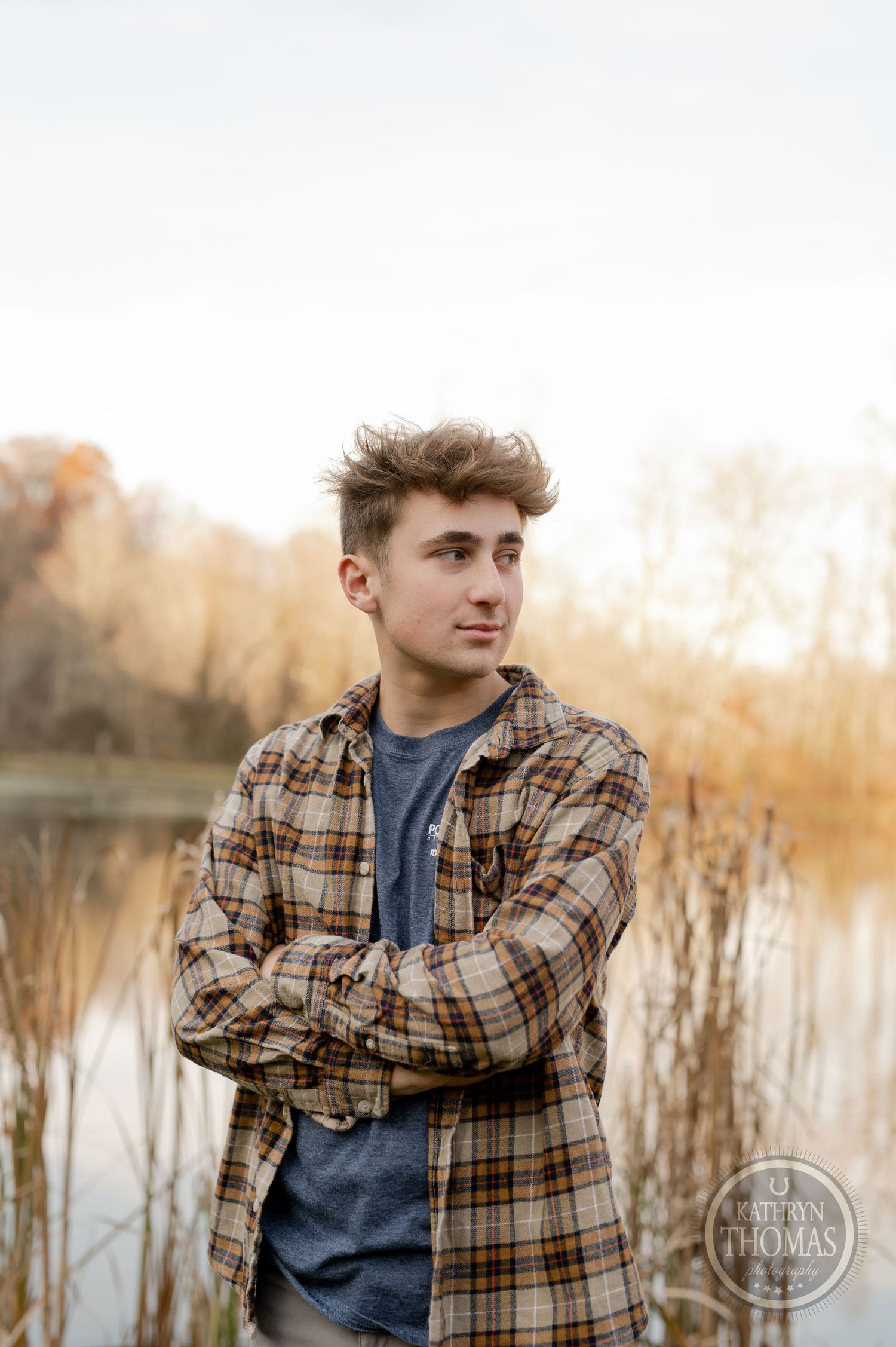 Someone wearing a brown plaid shirt stands near cattails and water with autumn trees in background.