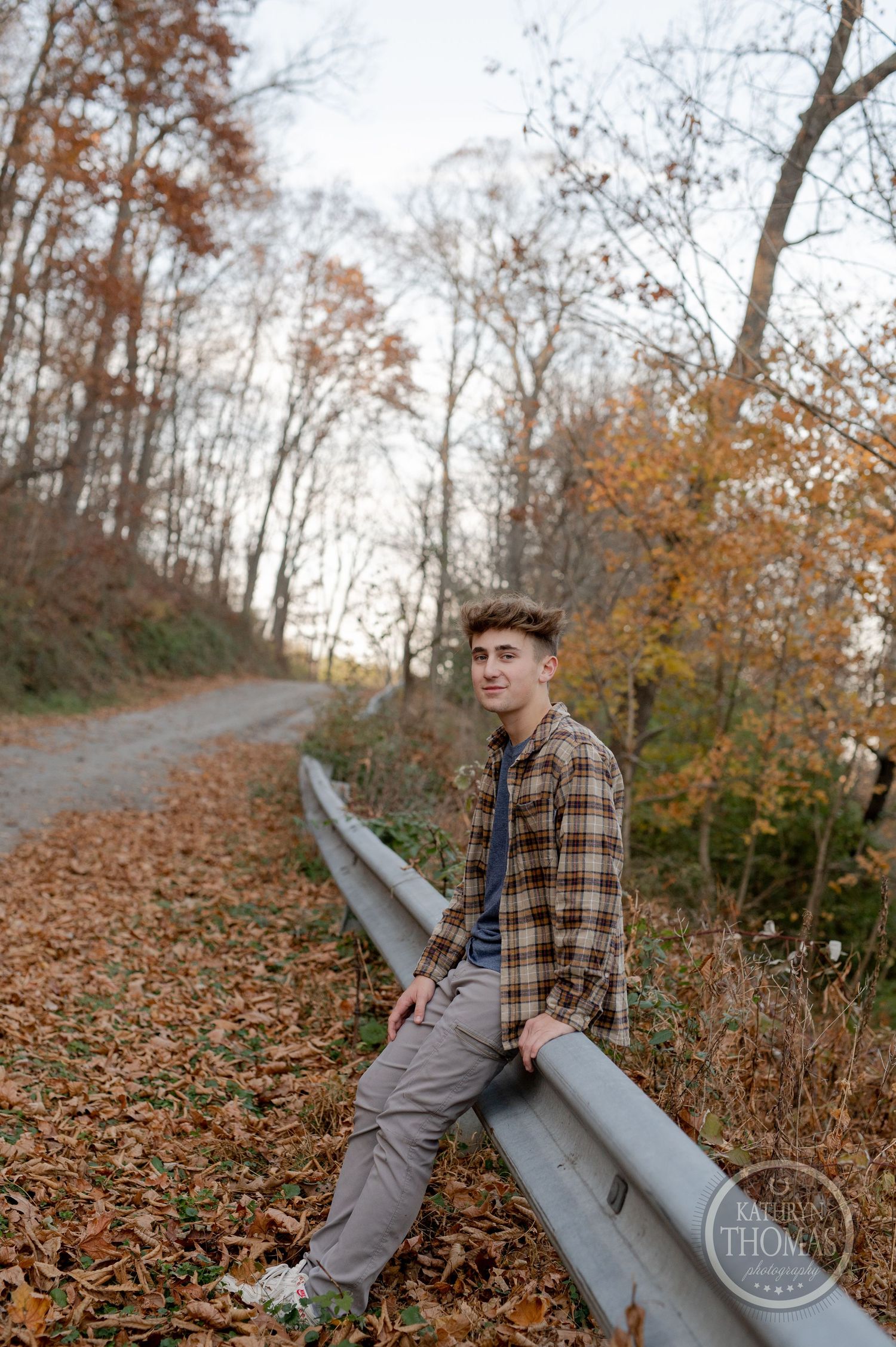 A person wearing a plaid flannel shirt sits on a guardrail along an autumn forest path covered in fallen leaves.