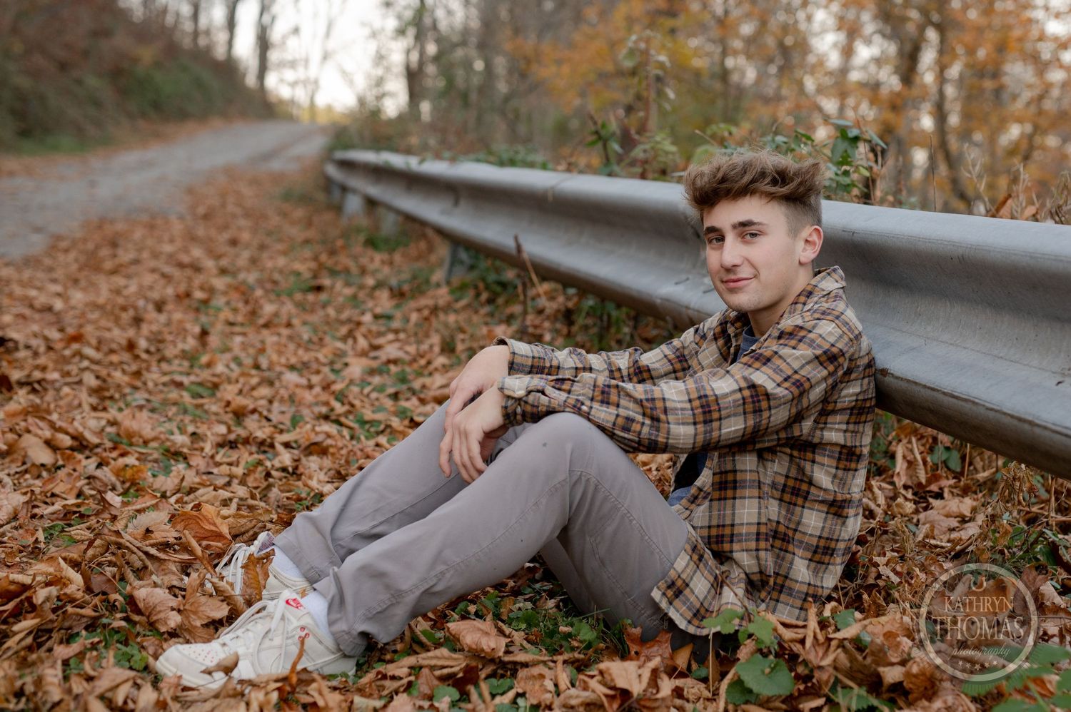 A casual portrait of someone sitting by a guardrail in autumn, wearing plaid and gray pants.