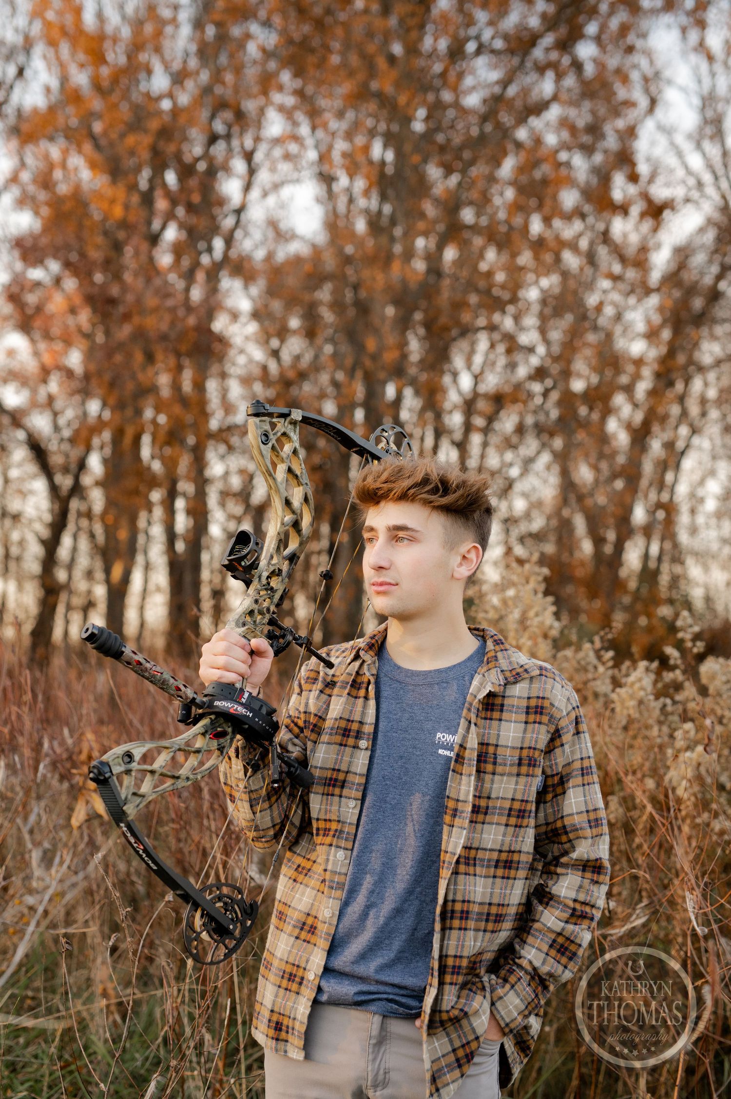 Archer in plaid shirt poses with camouflage compound bow in autumn field at sunset.