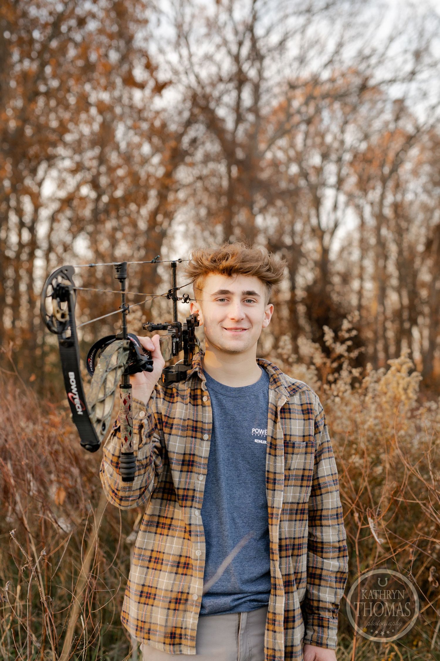 Archer smiles while holding compound bow in rustic autumn setting with brown foliage.