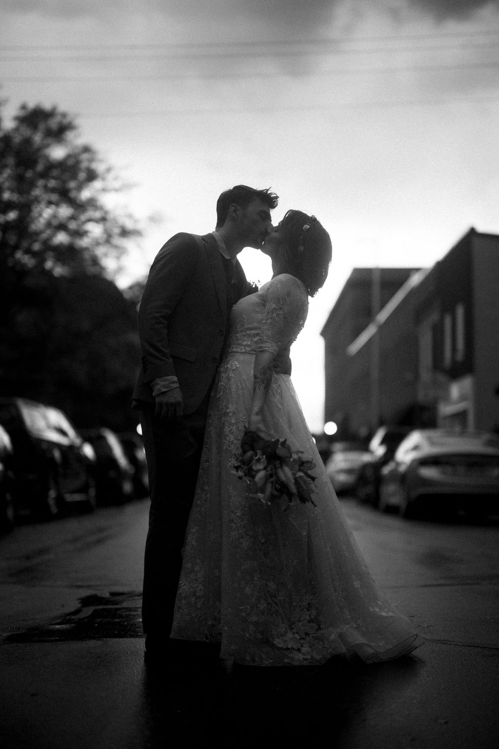 A romantic black and white silhouette of a bride and groom sharing a kiss on a city street at dusk.