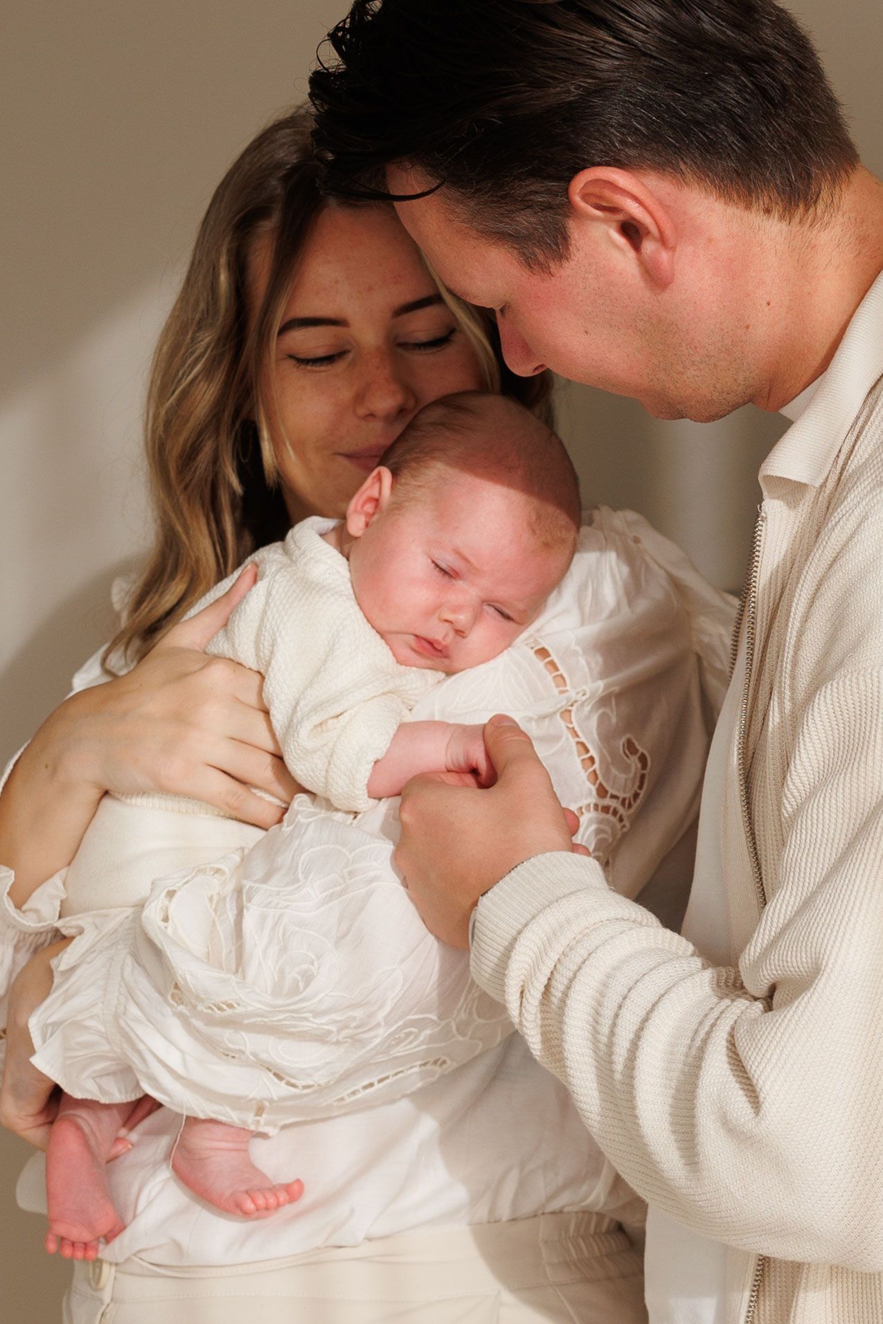A tender family moment as parents in white clothing gently embrace their sleeping newborn baby in a white dress.