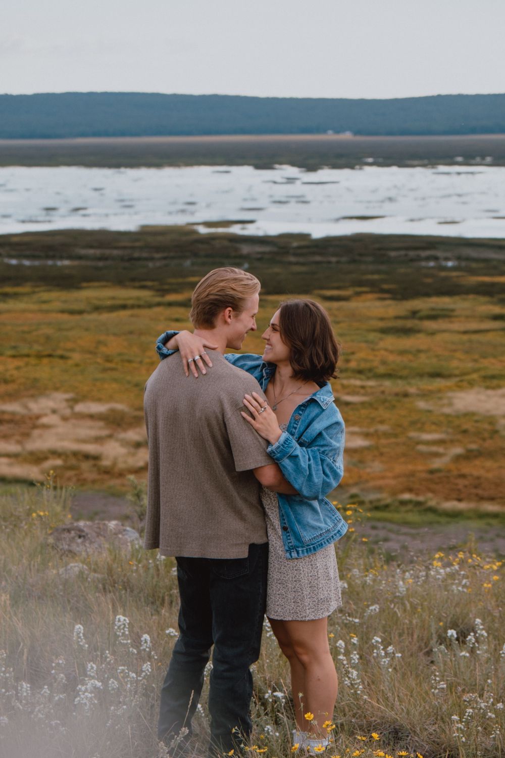 Two people share an intimate moment together in a wild meadow with mountains and water in the background.
