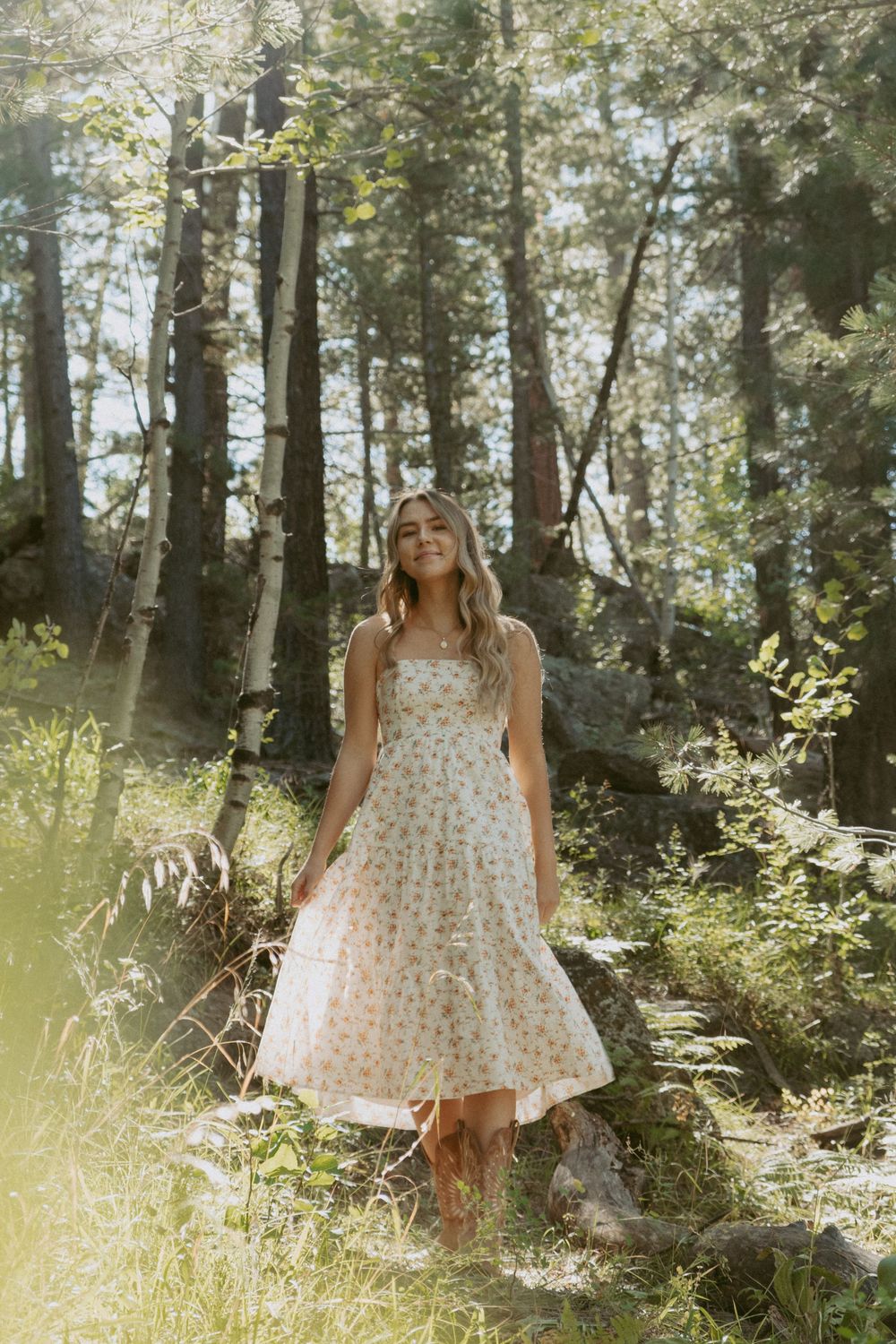 A person in a floral sundress stands in a sunlit forest clearing surrounded by tall trees and dappled sunlight.