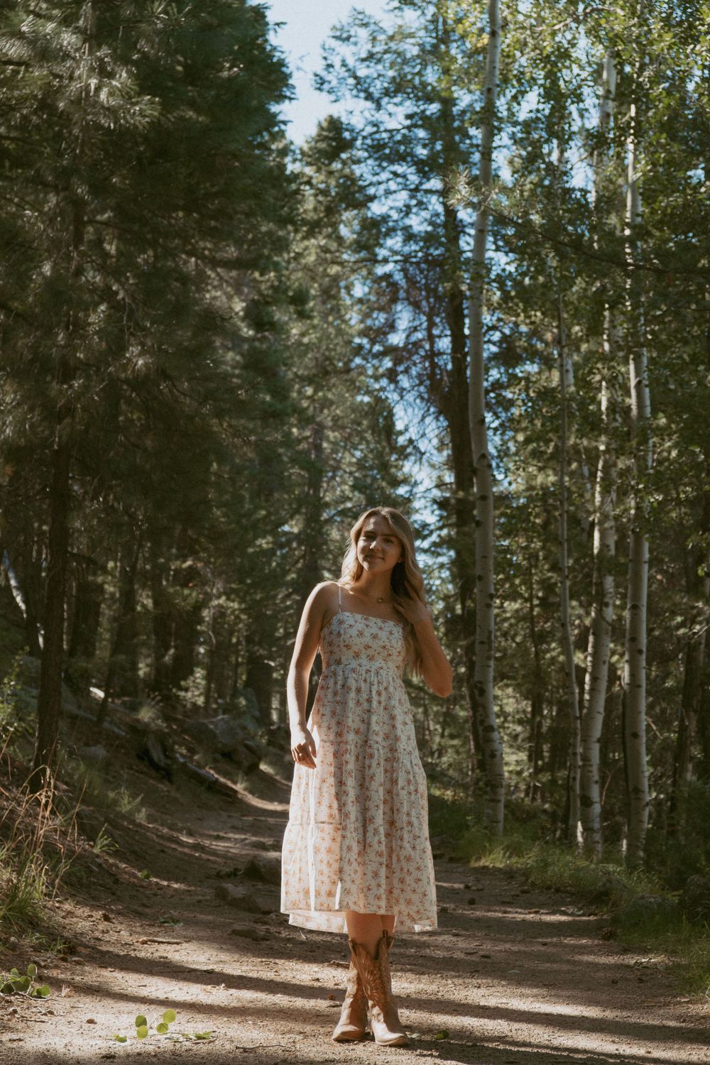 Figure in a floral summer dress and boots walks along a sunlit forest trail bordered by tall trees.