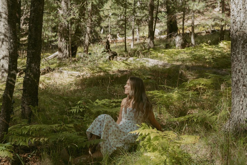 Someone in a floral dress sits on mossy ground in a sunlit forest while a deer grazes in the background.