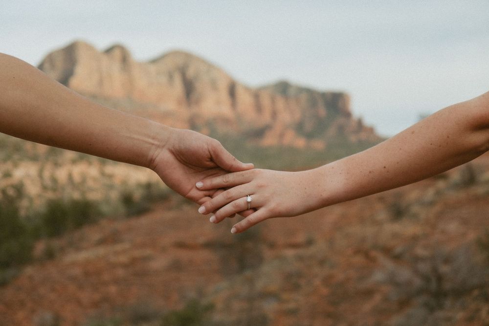 Two hands reaching to hold each other against a backdrop of red desert cliffs and mountains in warm sunlight.