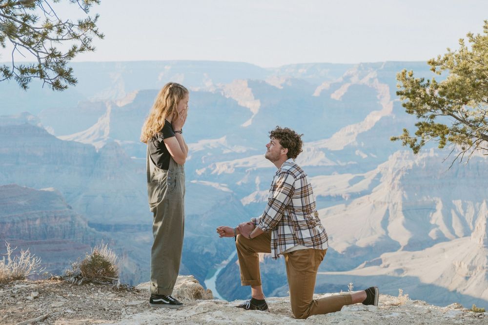 A couple shares a romantic proposal moment overlooking the scenic Grand Canyon landscape on a sunny day.