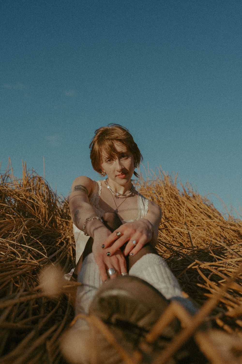 Person sitting in golden hay field on sunny day wearing casual clothing with moody vintage aesthetic.