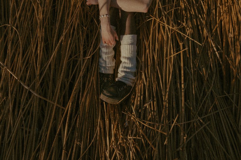 Legs wearing white knit socks and dark leather shoes rest against golden dried wheat stalks in moody lighting.