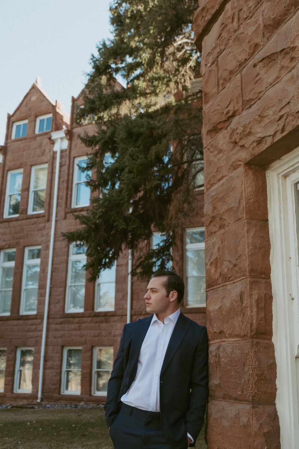 A person in a dark suit stands against a red brick building with classic architecture on an autumn day.